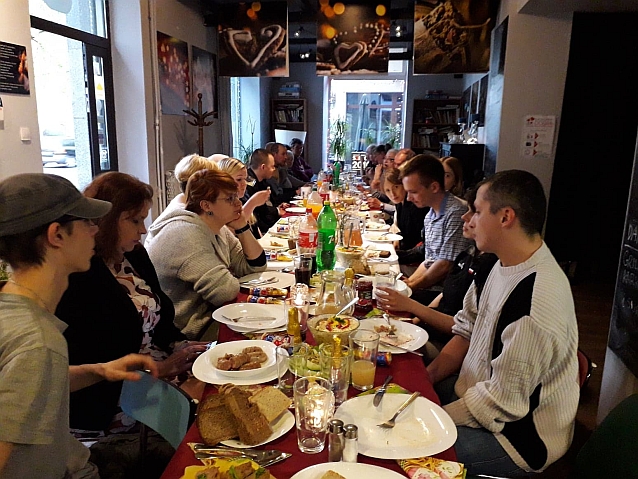 Ok. 20 osób siedzi po obu stronach długiego stołu, zastawionego jedzeniem i piciem