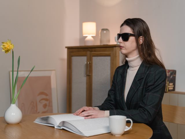 niewidoma dziewczyna w ciemnych okularach z długimi wlosami i marynarce siedzi przy stole w pokoju i czyta brajlem