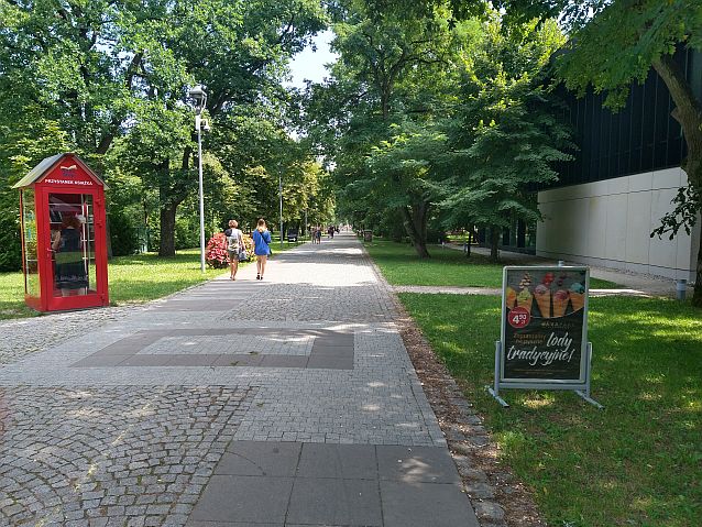 Szeroka alejka parkowa otoczona drzewami. Po lewej stronie czerwona budka, po prawej reklama lodziarni