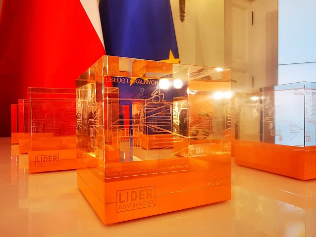Statuetki Konkursu Lider Dostępności 2021 stoją na stole. W tle flagi Polski i Unii Europejskiej