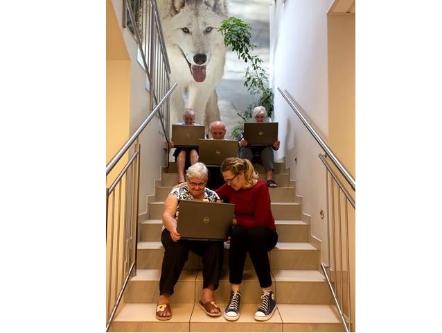 czwórka seniorów z laptopami na kolanach siedzą na schodach obok jednej z pań młoda dziewczyna