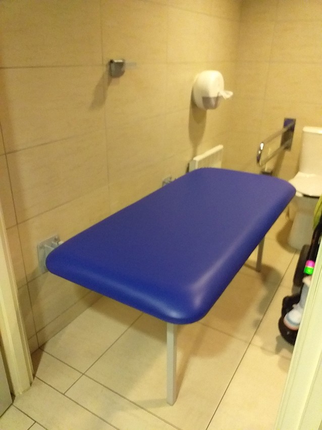 Przymocowana do ściany w toalecie rozłożona leżanka do przewijania dorosłych osób z niepełnosprawnością