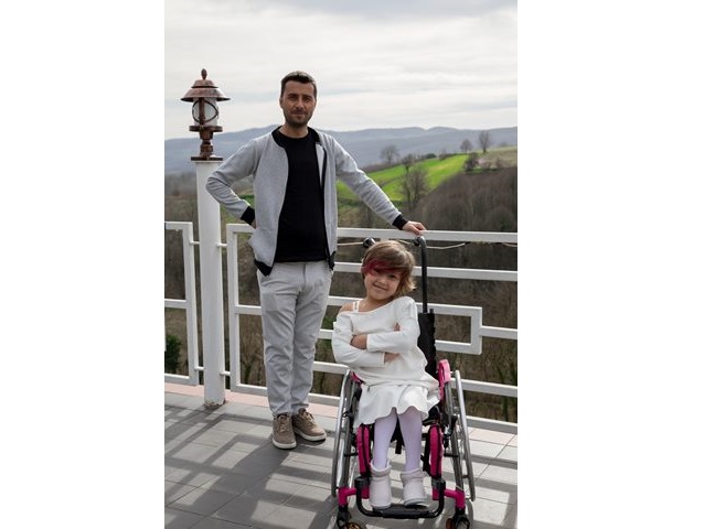 ojciec stoi na tarasie widokowym opierając jedną rękę na rączce wózka inwalidzkiego z dziewczynką