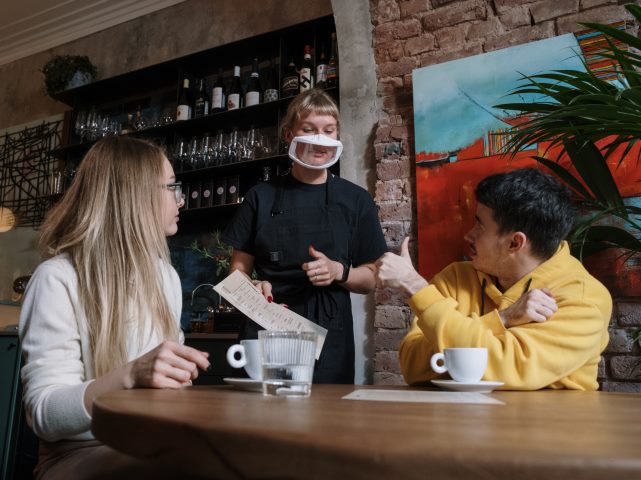 niesłysząca dziewczyna i chłopak w kawiarni obsługuje ich kelnerska w masce gdzie widać usta