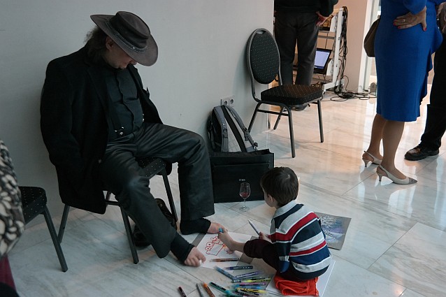 Stanisław Kmiecik maluje obraz stopami na podłodze. Naprzeciwko niego także stopami maluje mały chłopiec