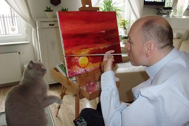 Piotr Pawłowski maluje obraz trzymając pędzel w ustach. Siedzący obok kot chce łapką trącić pędzel