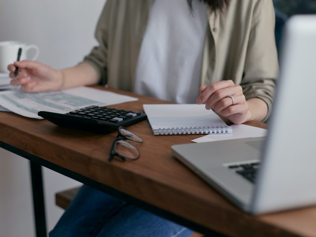 kobieta siedzi przy stole przed nią kartki kalkulator trzyma długopis w ręce stoi otwarty laptop