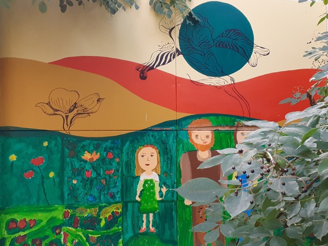 mural inspirowany twórczością S.Wyspiańskiego, prosty rysunek dziewczynki, kobiety i mężczyzny, w tle szerokie paski w kolorach: zielony, beżowy, czerwone kółko