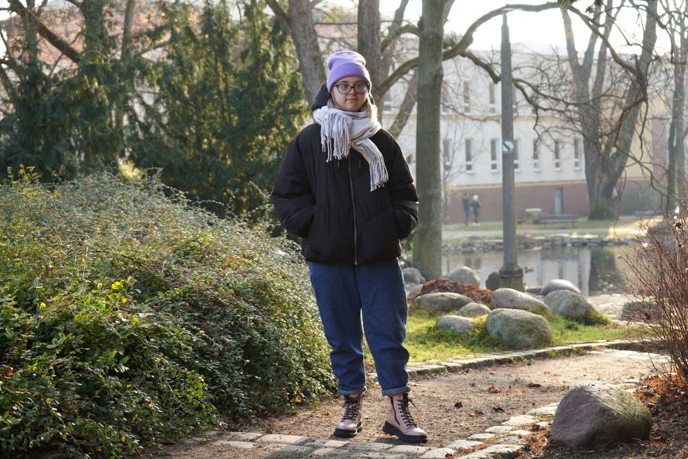 Paulina Rakowska ubrana w kurtkę i fioletową czapkę ma ręce w kieszeniach, stoi w parku