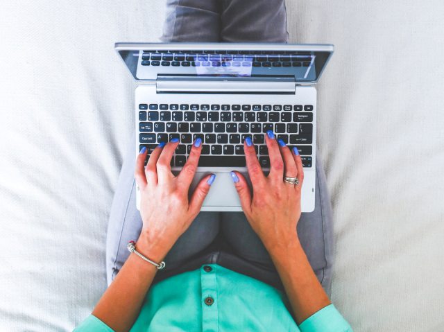 kobieta siedzi na łóżku, na kolanach ma laptopa, na którym pisze