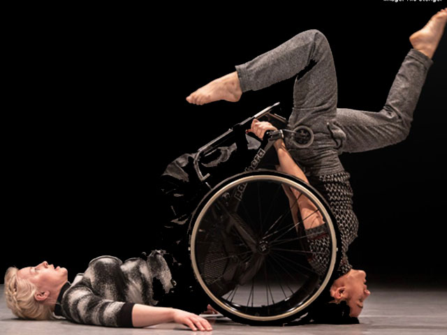 dwie osoby w tańcu. Kobieta na wózku leży plecami na podłodze, druga osoba stoi na głowie, trzyma się dłońmi o ramę wózka, 