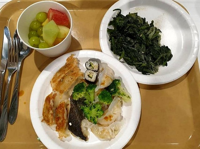 Taca z talerzami z jedzeniem, m.in. sushi, smażoną rybą, brokułami, wodorostami, a także owoce. Obok leżą sztućce