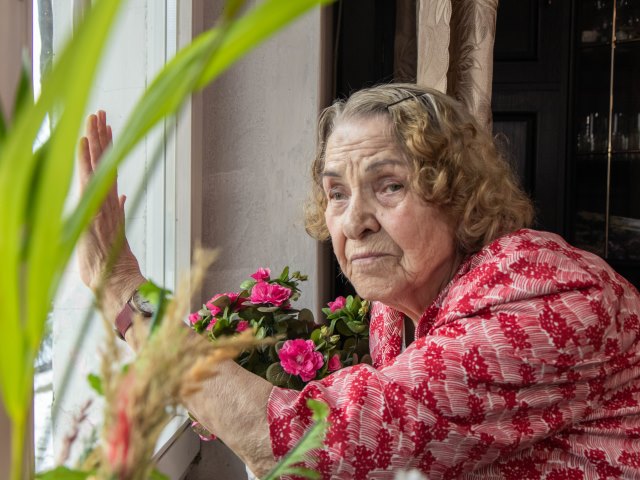 Pani Irena starsza kobieta z półdługimi włosami ze smutną miną stoi przy oknie na którym trzyma rękę