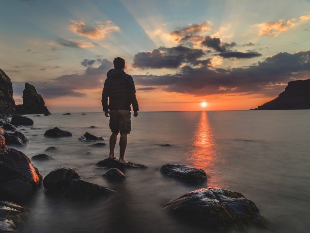 mężczyzna stoi na wystających z morza skałach, patrzy na zachód słońca
