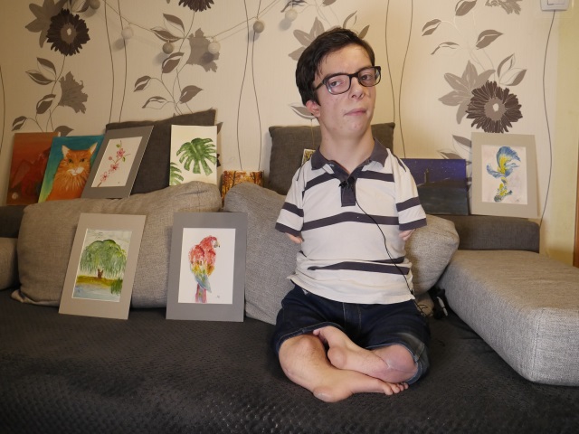 Milosz Bronisz, nastoletni chłopiec bez rąk za to w okularach siediz na kanapie. Obok rozłozone są jego rysunki.