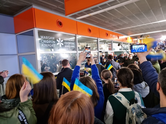 Tłum osób na hali przylotów na lotnisku, witający wracających z Pekinu ukraińskich paraolimpijczyków. Ludzie trzymają małe ukraińskie flagi