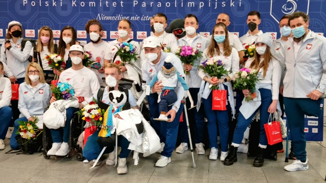 grupa rezprezentacji Polski pozuje do zdjęcia. Sportowcy mają maski na twarzach, są ubrani w białe koszulki, kurtki, bluzy z orłem Polski na piersi