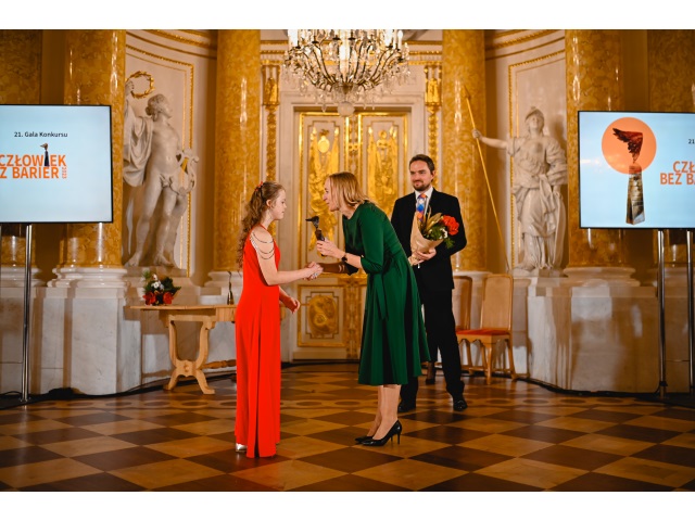 Ubrana w czerowną sukienę Wanessa Bąkowska, młoda dziewczyna z zespołem Downa odbiera nagrodę z rąk ubranej w zieloną suknię kobiety na Sali Balowej Zamku Królewskiego w Warszawie. 