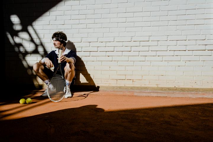 zdjęcie poglądowe. Mężczyzna siedzi pod ścianą. W dłoni trzyma rakietkę do tenisa. 