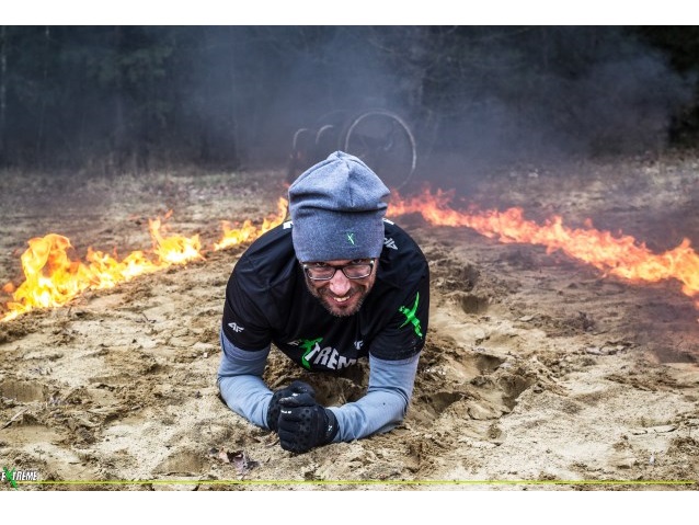Mężczyzna w czapce i okularach czołga się po piasku bez wózka, z dwóch stron są linie z ognia|