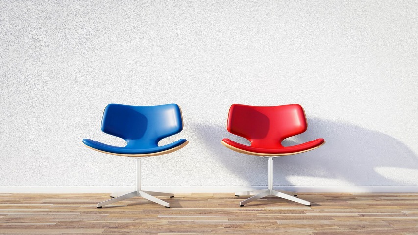 Zdjęcie przedstawia dwa puste krzesła na tle jasnej ściany. Krzesła wyróżnia to, że jedno jest czerwone a drugie niebieskie. Dzięki czemu kontrast jest jeszcze bardziej widoczny.