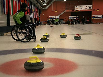 Osoba siedząca na wózku, który stoi na lodzie, obok leżą kamienie do curlingu.