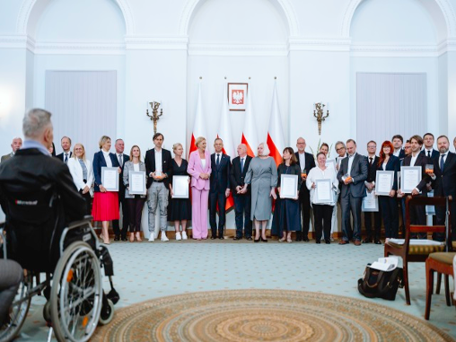 Nagrodzeni i wyróżnieni w konkursie Lider dostępności stoją razem z organizatorami i prezydentem w Sali w Pałacu Prezydenckim. Przed nimi jest mężczyzna na wózku.